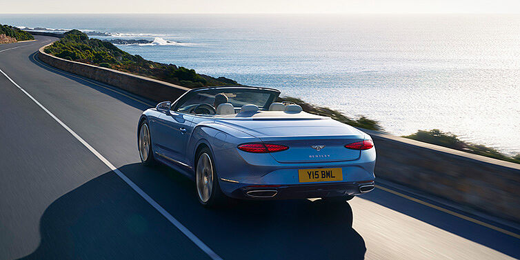 Bentley Vilnius New Bentley Continental GTC Azure convertible rear view in Light Windsor blue paint with 22 inch ten spoke wheels driving dynamically on an ocean road in the sunshine