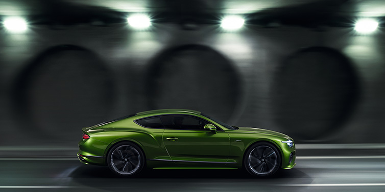 Bentley Vilnius Bentley Continental GT Speed coupe profile in Tourmaline Green driving dynamically past a concrete wall at night
